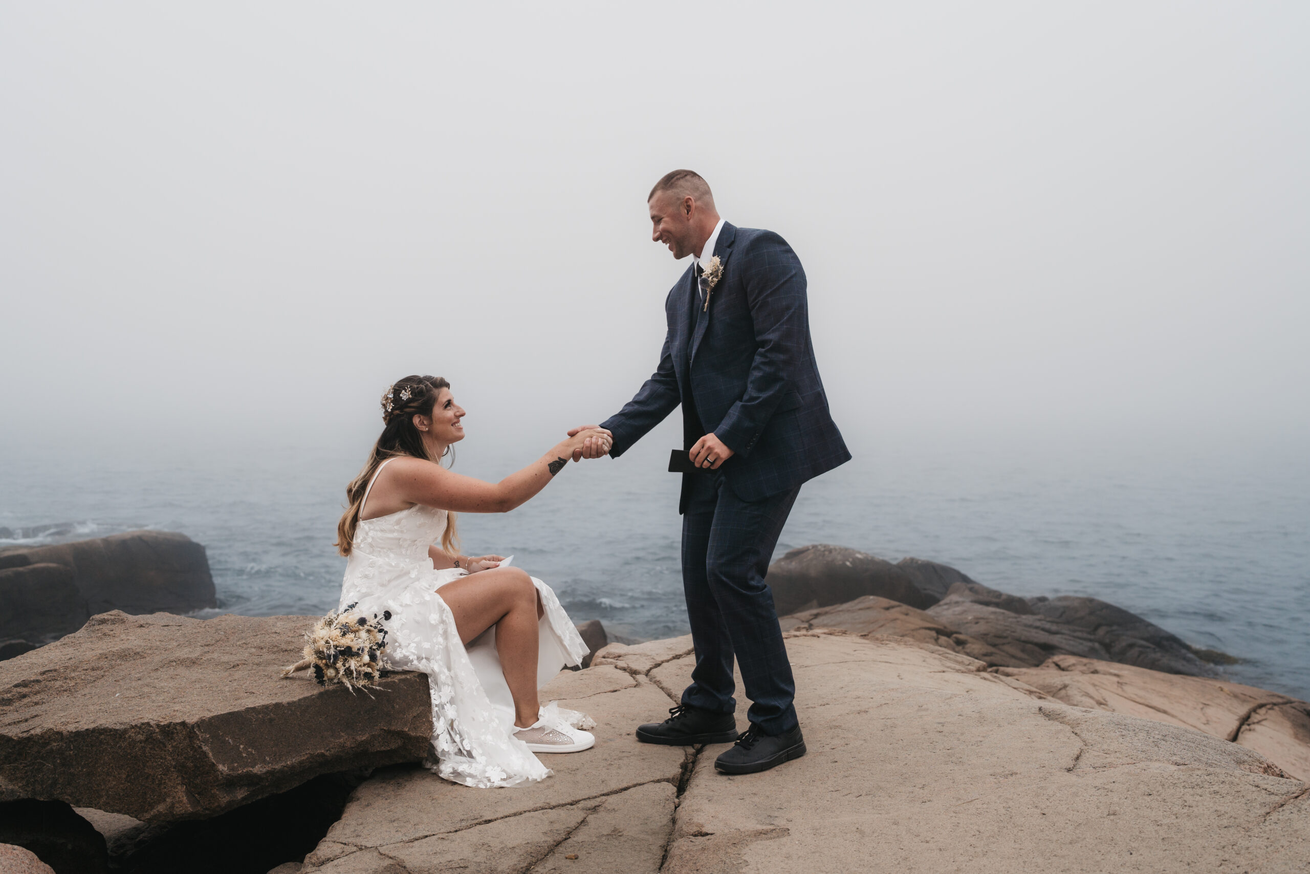 Groom helping his bride up on the rocky coat of Otter point.
